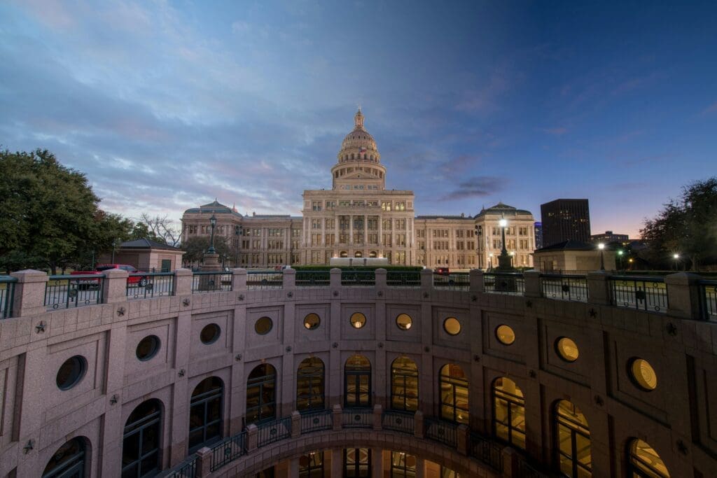 The Texas State Capitol building in Austin is illuminated at dusk, with a cloudy sky overhead and circular lower-level windows visible in the foreground.