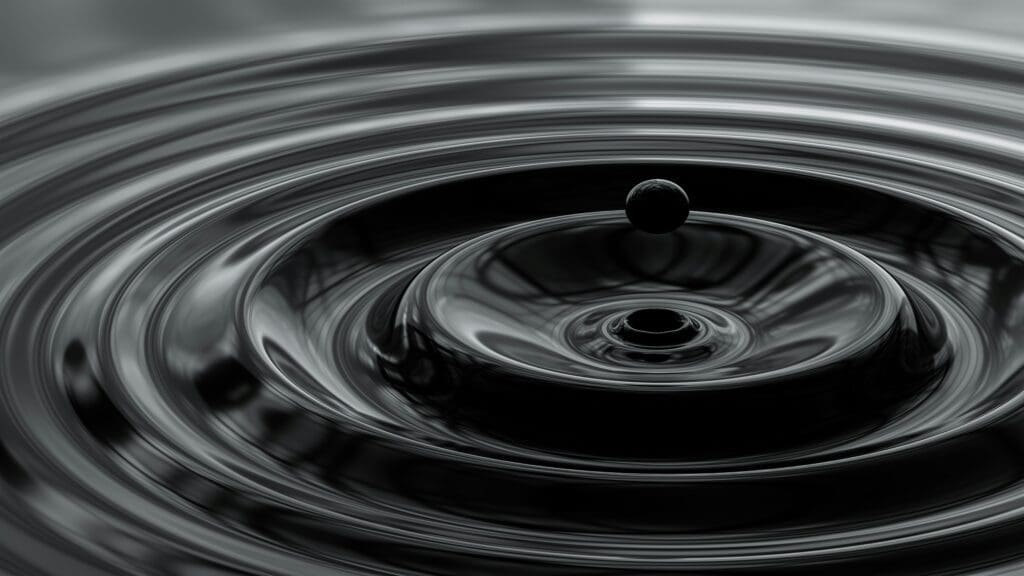 A close-up of a single water droplet suspended above the surface, creating circular ripples in the dark, reflective liquid below.