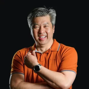 A middle-aged man with gray hair wearing an orange polo shirt and a smartwatch, smiling with his arms crossed against a plain black background.