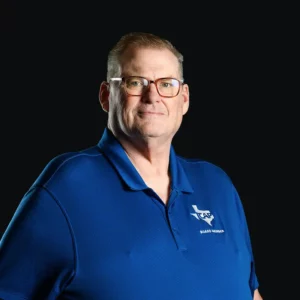 A middle-aged man with short hair and glasses wears a blue collared shirt with a Texas logo and "Board Member" text, standing against a plain black background.