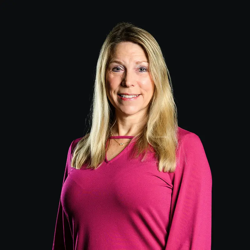 A woman with straight, blonde hair wearing a long-sleeved, pink top stands smiling against a solid black background.