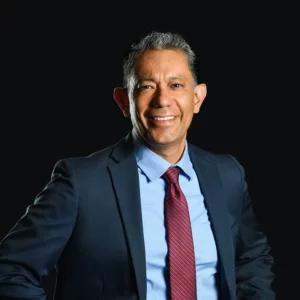 A man in a dark suit, light blue shirt, and red tie stands smiling with hands on hips against a plain black background.