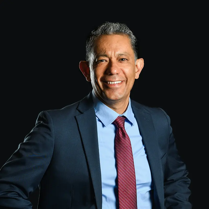 A man in a dark suit, light blue shirt, and red tie stands smiling with hands on hips against a plain black background.