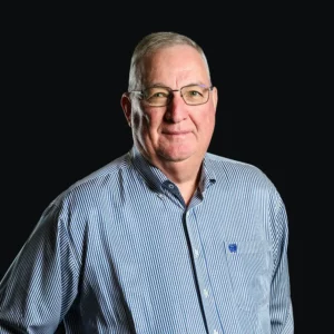 An older man with short gray hair and glasses, wearing a blue and white striped button-up shirt, stands against a plain black background, looking at the camera with a neutral expression.