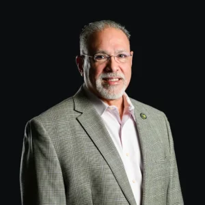 A man with gray hair and a beard, wearing glasses, a light pink shirt, and a gray checkered blazer, poses against a solid black background. He is smiling and wearing a round lapel pin.