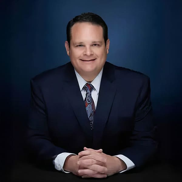 A man in a dark suit and patterned tie sits with his hands folded on a table, smiling, against a dark blue gradient background.