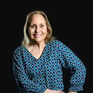 A smiling older woman with long blonde hair, wearing a blue patterned blouse, stands against a solid black background with one hand on her hip.