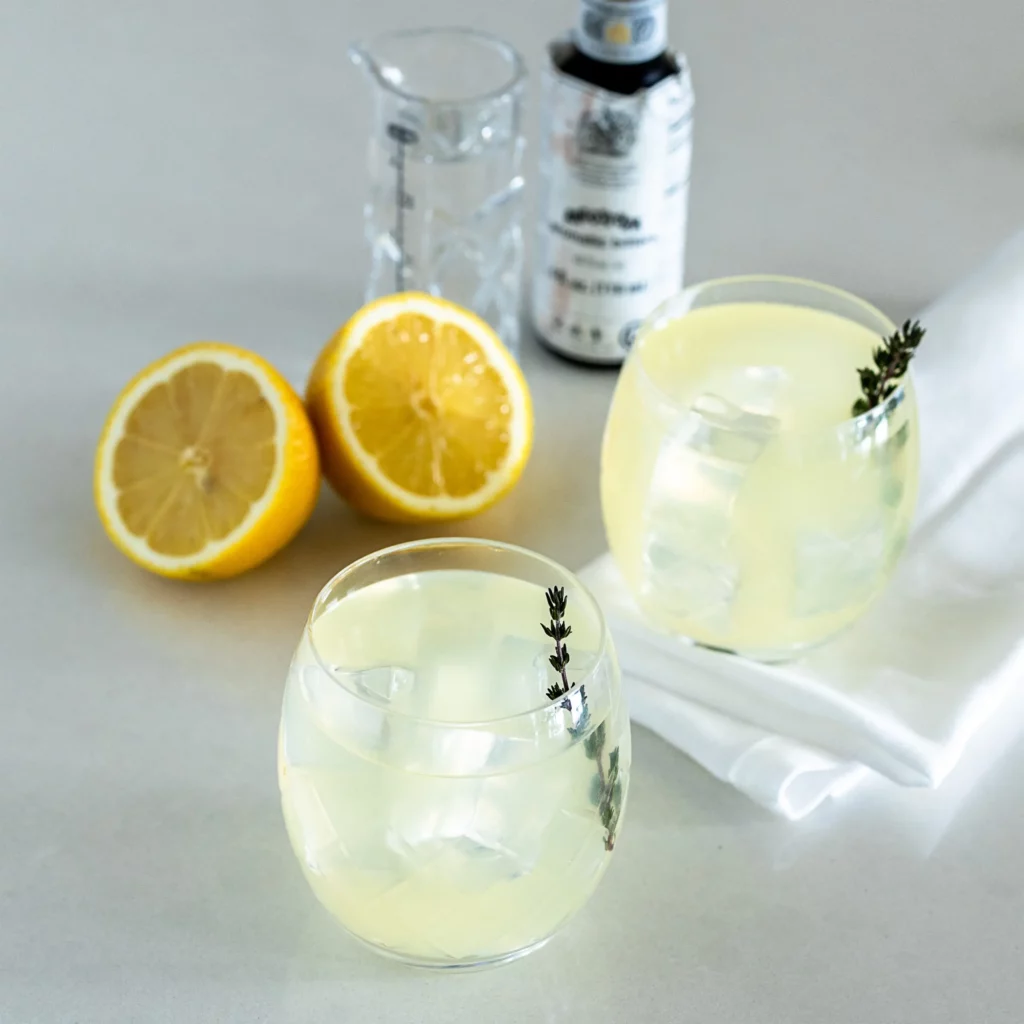 Two round glasses with a pale yellow drink, ice cubes, and a sprig of thyme. Next to them are a halved lemon, a clear measuring glass, a bottle, and a white napkin on a light countertop.
