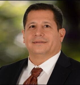 A man wearing a dark suit, white shirt, and patterned brown tie smiles at the camera. He has short dark hair and is pictured against a blurred outdoor background with green and yellow tones.