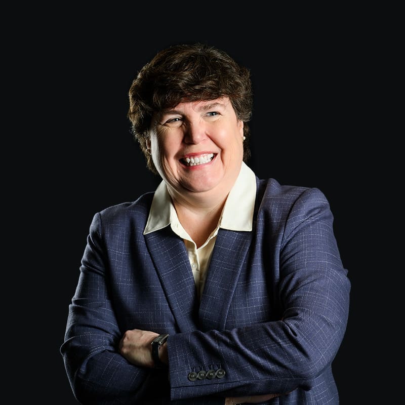 A person with short brown hair, wearing a blue blazer and white collared shirt, smiles with arms crossed in front of a plain black background.