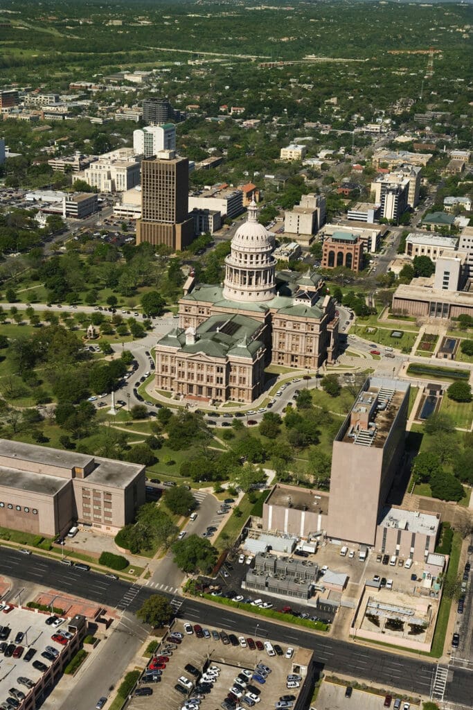 Aerial view of the Texas State Capitol building surrounded by trees and downtown Austin, with streets, parked cars, and city buildings visible in the background.