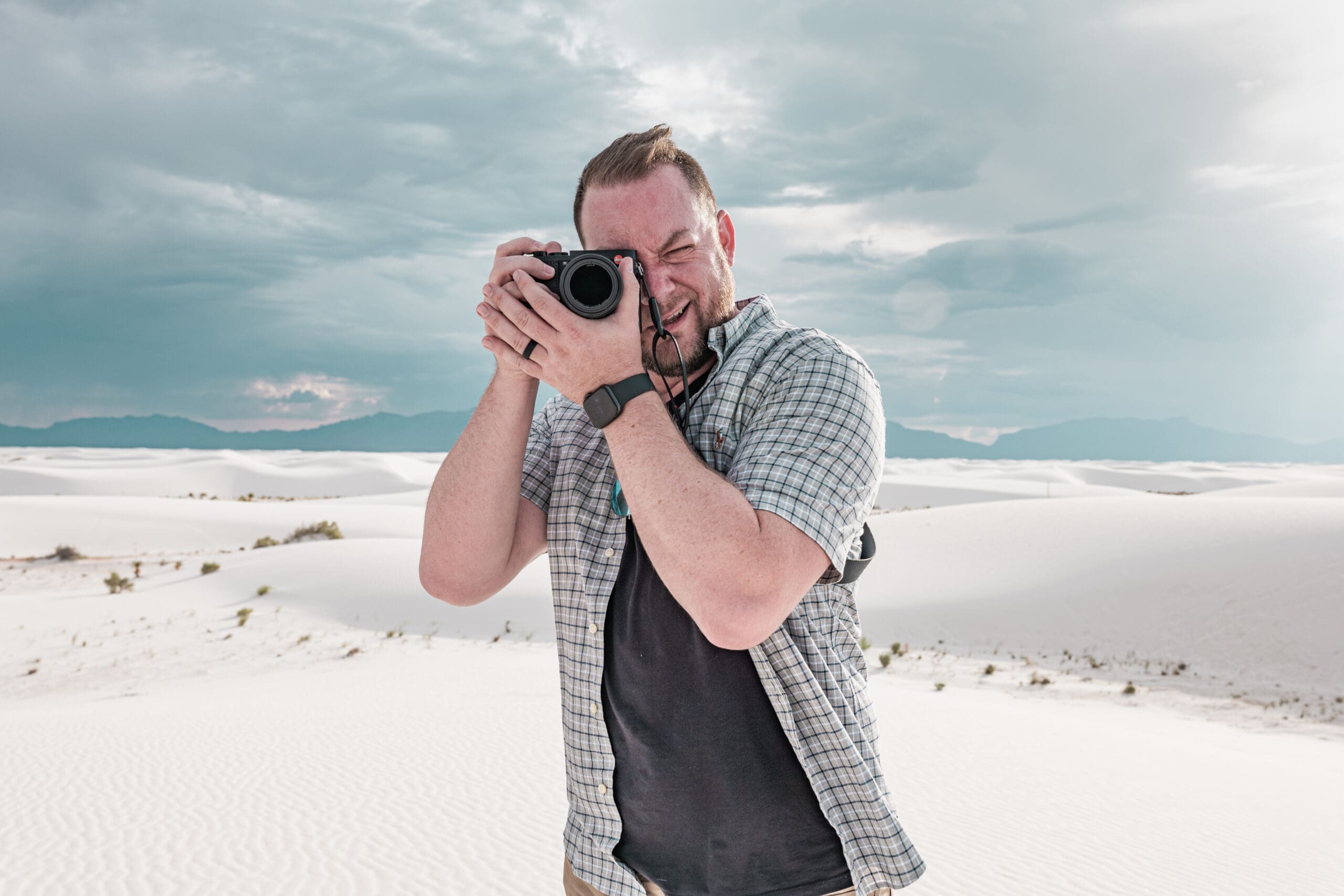 A person standing in a white sand desert, wearing a short-sleeve plaid shirt and holding a camera up to their face, taking a photo with mountains and cloudy sky in the background.