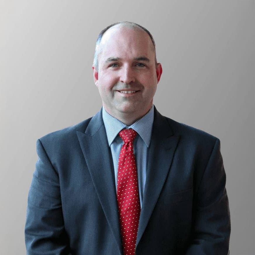A man with short hair, wearing a dark suit, light blue shirt, and red polka dot tie, smiles while sitting against a plain, light gray background.
