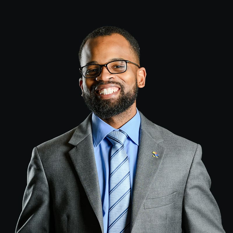 A man with a trimmed beard and glasses smiles, wearing a gray suit jacket, a light blue shirt, and a striped tie, against a plain black background.